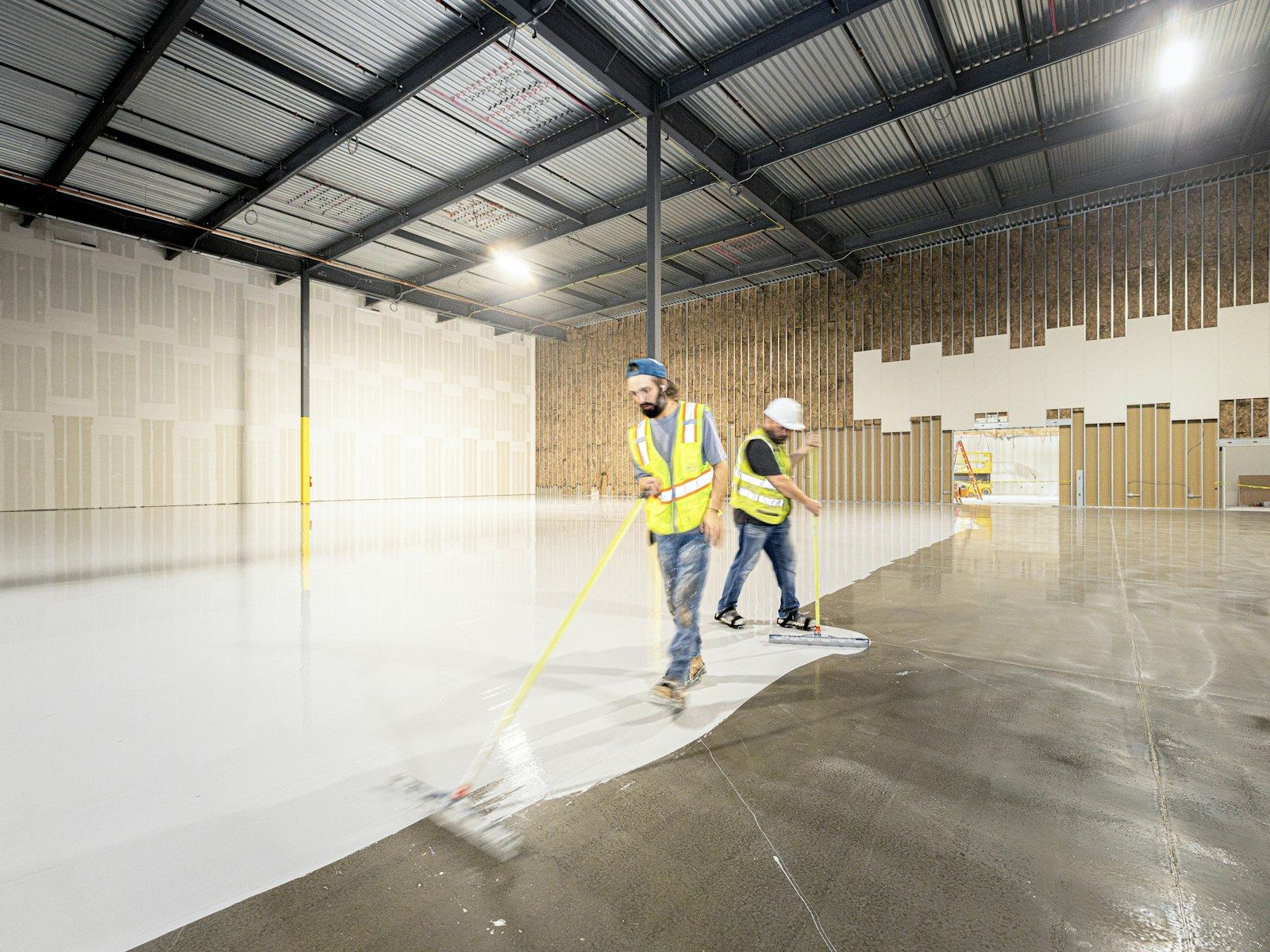 Two workers in hi-vis spreading white epoxy coating across a warehouse floor.
